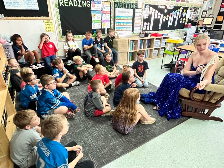 girl in blue dress reading to class of kids